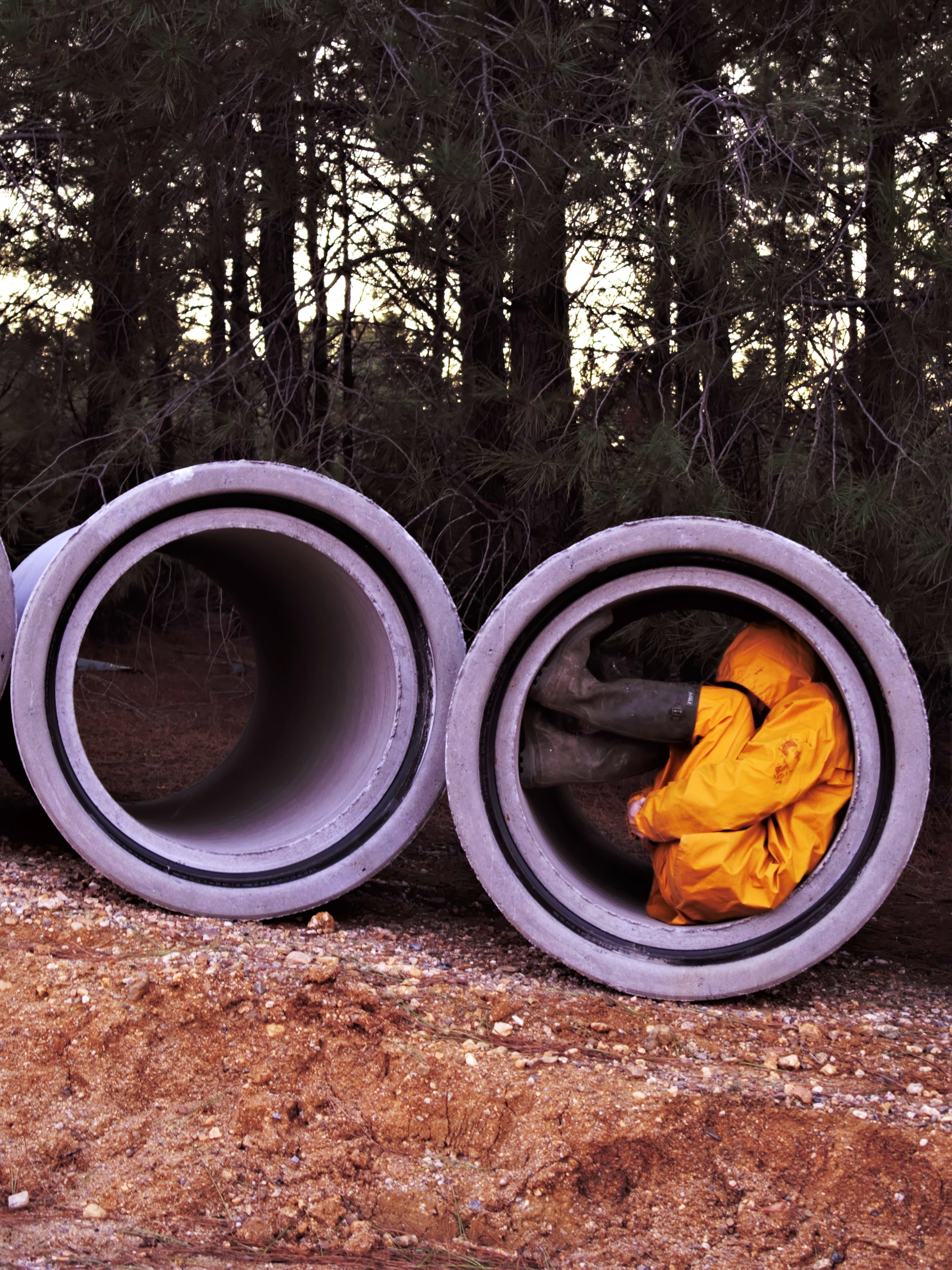 A person in a yellow raincoat and rain boots curled up in a concrete pipe